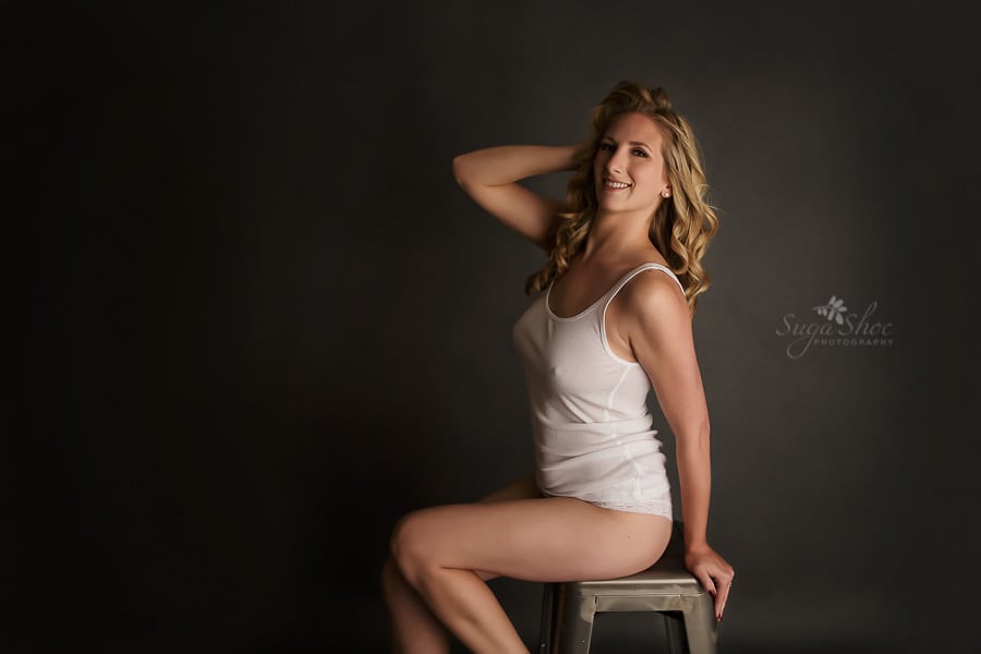 SugaShoc Photography Bucks County Boudoir Photographer PA boudoir pose sitting on stool wearing white tank top and panty with hand in hair smiling