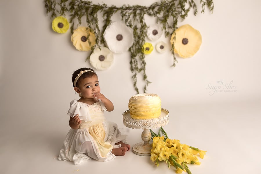 SugaShoc_Photography_Cake_Smash_Photographer_Bucks_County_PA_Doylestown_PA_Cream_and_yellow_theme_tasting_frosting_cake_on_hands_yellow_flowers