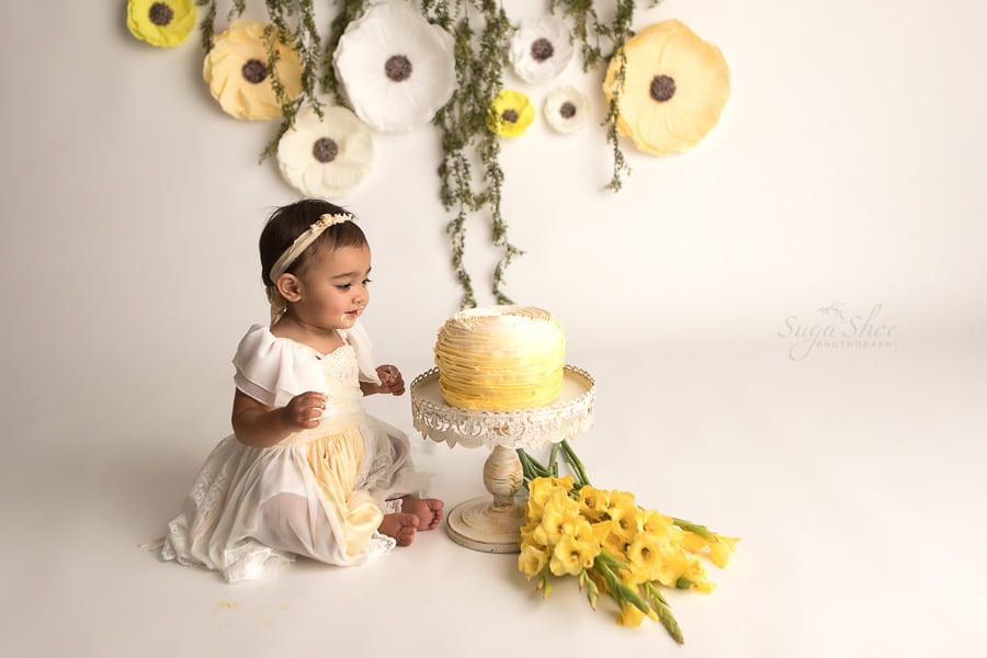 SugaShoc_Photography_Cake_Smash_Photographer_Bucks_County_PA_Doylestown_PA_Cream_and_yellow_theme_staring_at_cake_cake_on_hands_yellow_Flowers