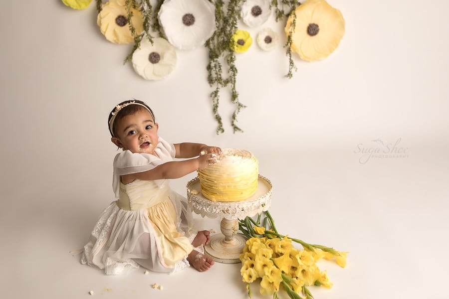 SugaShoc_Photography_Cake_Smash_Photographer_Bucks_County_PA_Doylestown_PA_Cream_and_yellow_theme_looking_at_camera_cake_on_hands_yellow_Flowers