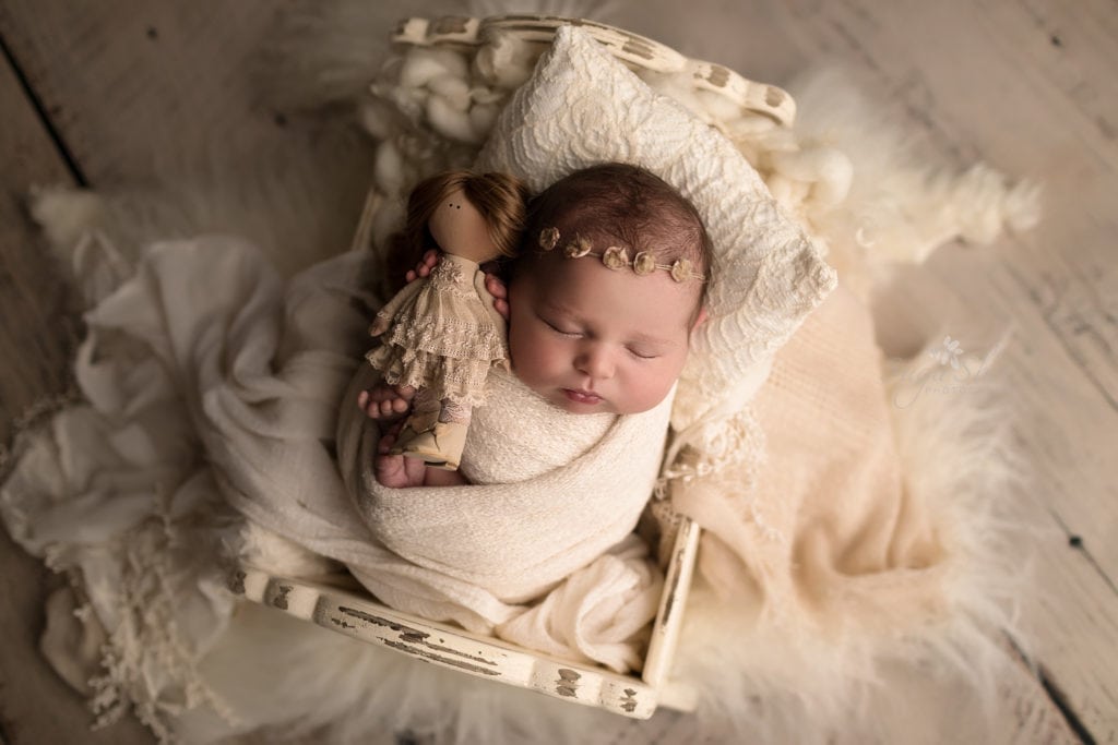SugaShoc Photography Newborn Photographer Bucks County PA Doylestown PA Neutral Tones Baby girl sleeping on white antique bed wrapped in cream wrap with golden floral headband holding a dolly with other cream colored wraps and throws