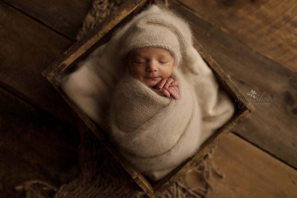 SugaShoc Photography Newborn Photographer Bucks County PA Doylestown PA Neutral Tones Baby wearing cream knit hat sleeping in antique wooden crate wrapped in cream knit wrap on wooden floor