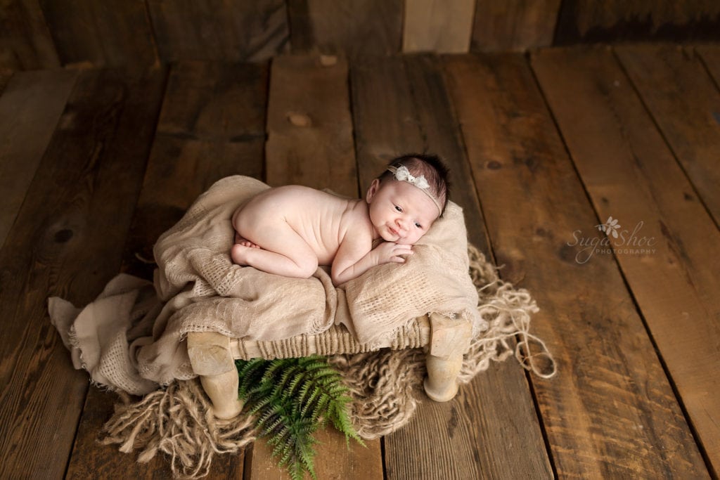 SugaShoc Photography Newborn Photographer Bucks County PA Doylestown PA Keeping Babies Asleep Baby girl awake laying on wooden stool with tan blanket and wearing bow headband