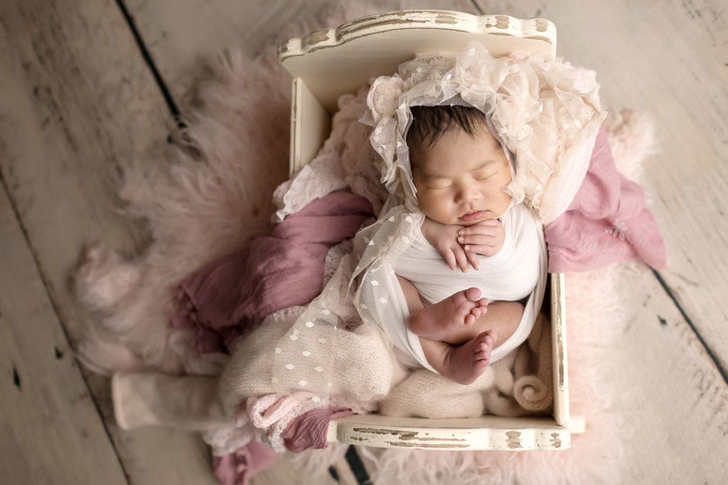 SugaShoc Photography Newborn Photographer Bucks County PA Doylestown PA Sophia's Newborn Session Baby girl sleeping in white antique bed with pink and purple throws and pale pink lace hat
