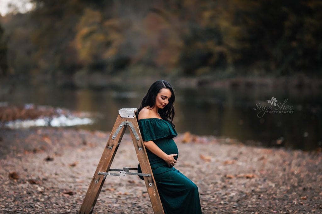 SugaShoc Photography Maternity Photographer Bucks County PA Doylestown PA Tyler State Park Maternity Session pregnant woman wearing deep green off the shoulder maternity dress leaning on a wooden ladder looking down at belly by river