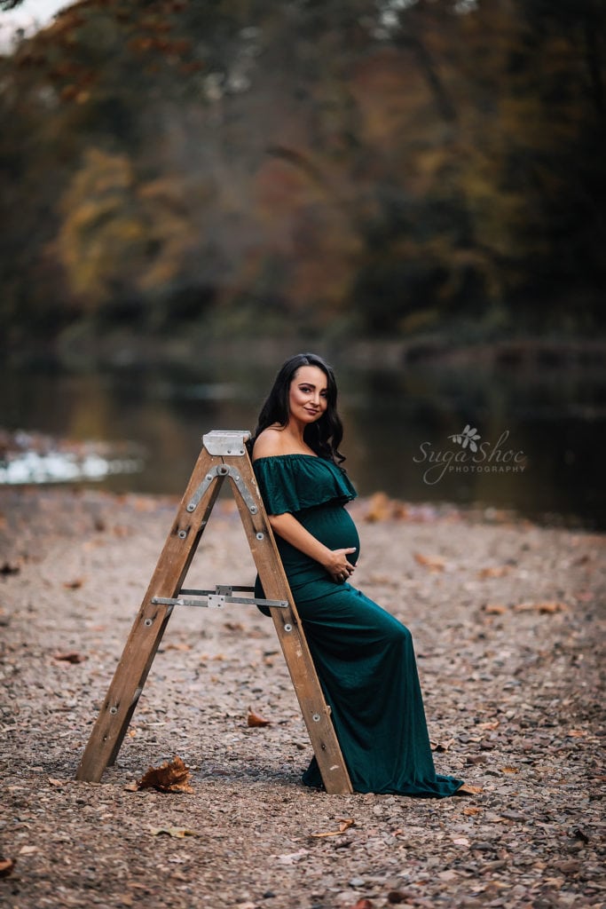 SugaShoc Photography Maternity Photographer Bucks County PA Doylestown PA Tyler State Park Maternity Session pregnant woman wearing deep green off the shoulder maternity dress leaning on a wooden ladder by river