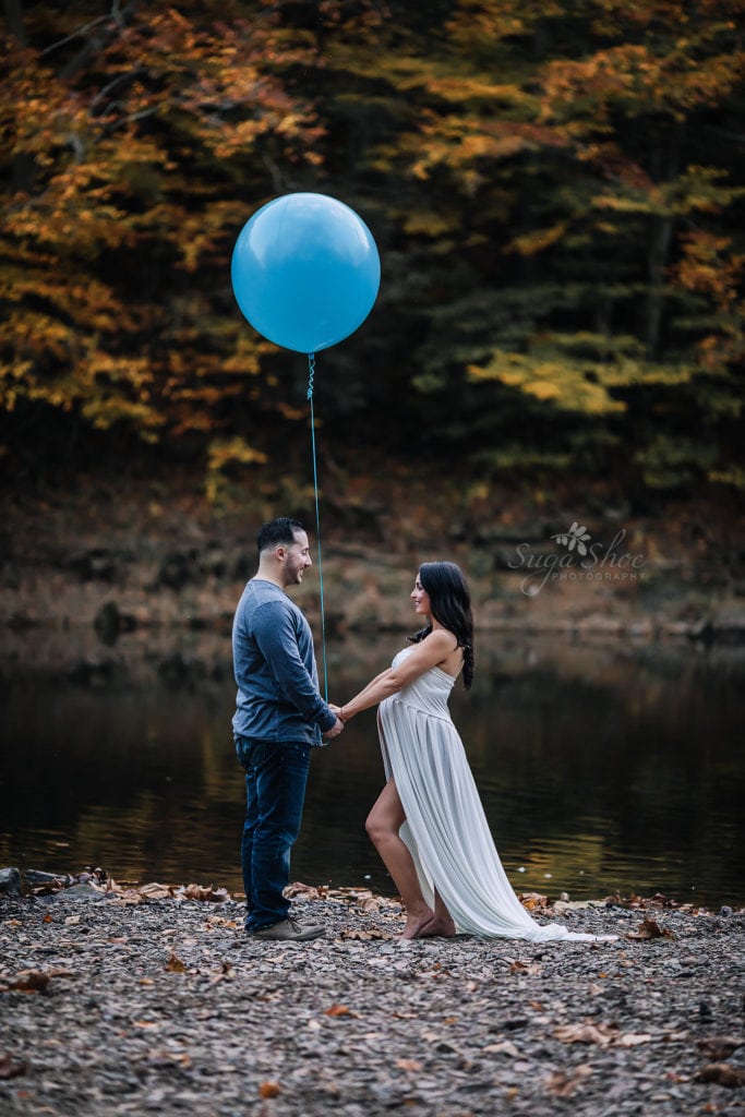 SugaShoc Photography Maternity Photographer Bucks County PA Doylestown PA Tyler State Park Maternity Session couple standing by river bed holding hands and bog blue balloon woman wearing white maternity dress