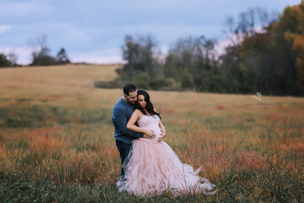 SugaShoc Photography Maternity Photographer Bucks County PA Doylestown PA Tyler State Park Maternity Session couple standing in field hands on pregnant belly woman wearing pink tulle maternity dress