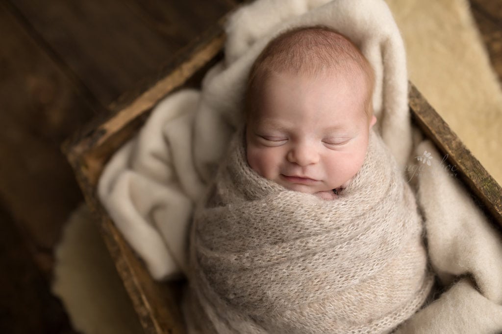 SugaShoc Photography Newborn Photographer Bucks County PA Doylestown PA Baby Hunter Newborn Session sleeping in neutral knit wrap in wooden crate with slight smile