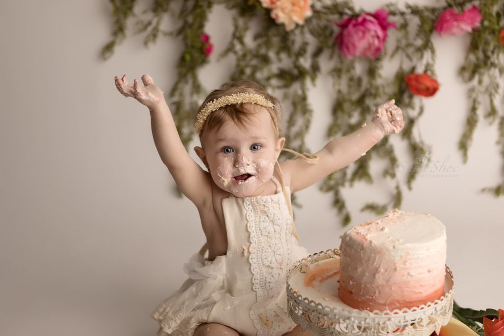 SugaShoc Photography Baby Photographer Bucks County PA Doylestown PA Cake Smash Session with Flowers baby arms in the air smiling by ombre cake in cream lace romper with floral garland and fresh flower bouquet boho chic 