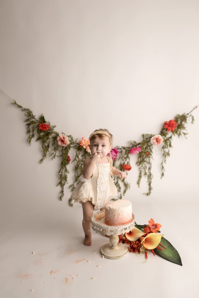 SugaShoc Photography Baby Photographer Bucks County PA Doylestown PA Cake Smash Session with Flowers baby standing by ombre cake in cream lace romper with floral garland and fresh flower bouquet boho chic 