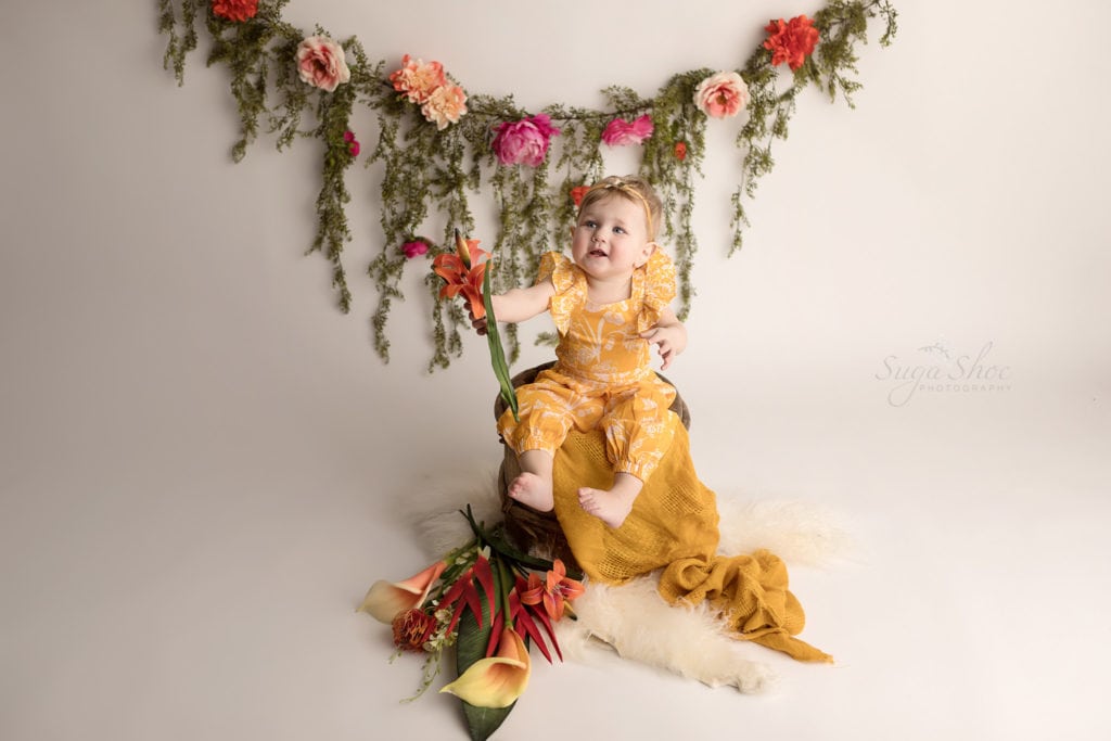 SugaShoc Photography Baby Photographer Bucks County PA Doylestown PA Cake Smash Session with Flowers baby wearing yellow floral romper sitting wooden barrel with yellow blanket and floral garland and holding fresh flower bouquet