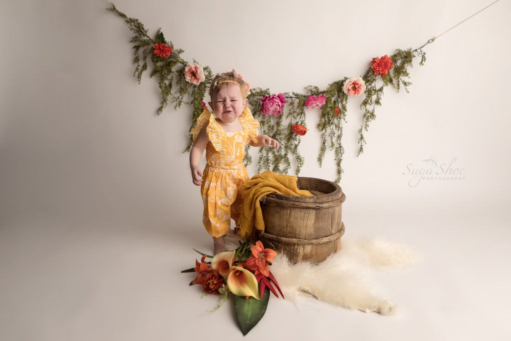 SugaShoc Photography Baby Photographer Bucks County PA Doylestown PA Cake Smash Session with Flowers baby wearing yellow floral romper crying standing next to wooden barrel with yellow blanket and floral garland and fresh flower bouquet