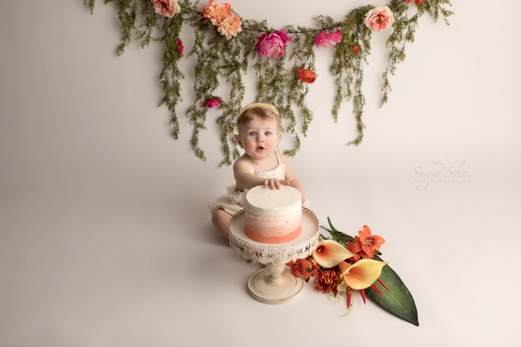 SugaShoc Photography Baby Photographer Bucks County PA Doylestown PA Cake Smash Session with Flowers baby touching ombre cake in cream lace romper with floral garland and fresh flower bouquet boho chic