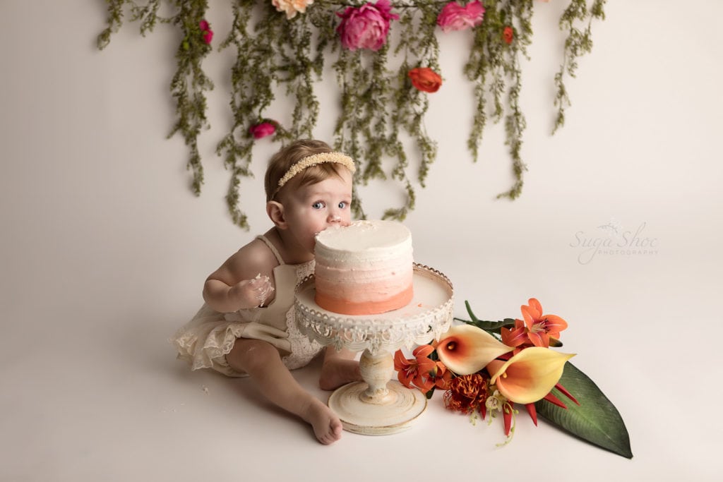 SugaShoc Photography Baby Photographer Bucks County PA Doylestown PA Cake Smash Session with Flowers baby eating ombre cake in cream lace romper with floral garland and fresh flower bouquet boho chic