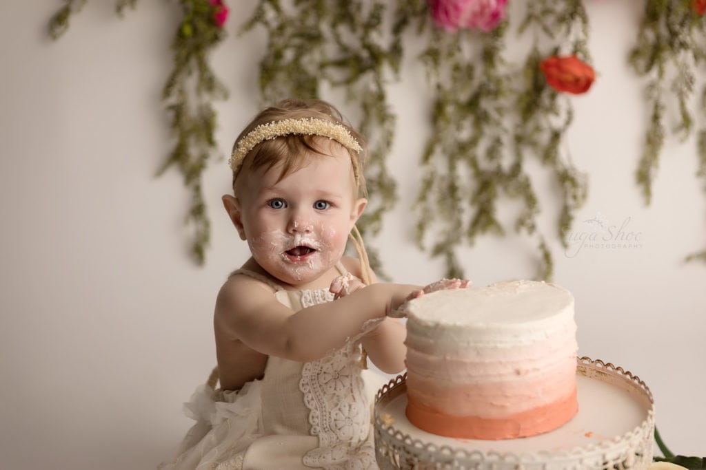 SugaShoc Photography Baby Photographer Bucks County PA Doylestown PA Cake Smash Session with Flowers baby touching ombre cake in cream lace romper with floral garland and fresh flower bouquet boho chic close-up