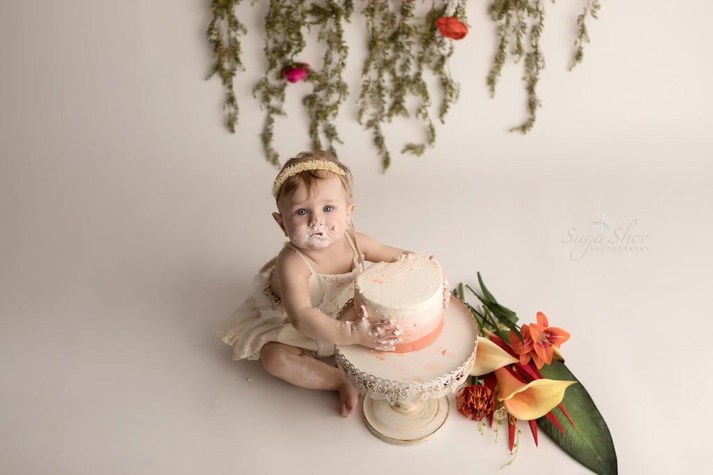 SugaShoc Photography Baby Photographer Bucks County PA Doylestown PA Cake Smash Session with Flowers baby holding ombre cake in cream lace romper with floral garland and fresh flower bouquet boho chic 