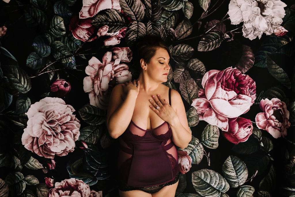 SugaShoc Photography Doylestown Boudoir Photographer PA Floral Boudoir Session boudoir pose standing against floral backdrop wearing dark red teddy with one hand on shoulder and one on clavicle looking off to side