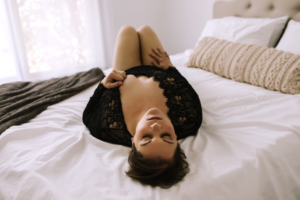 SugaShoc Photography Doylestown Boudoir Photographer PA Floral Boudoir Session boudoir pose laying on bed on back wearing blank lace bodysuit with one hand on thigh and one on breast