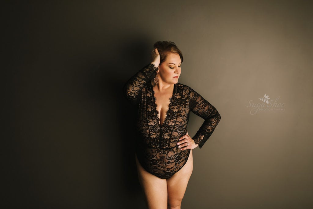 SugaShoc Photography Doylestown Boudoir Photographer PA Floral Boudoir Session boudoir pose standing in front on dark wall wearing black lace bodysuit with hands on head and hip looking down