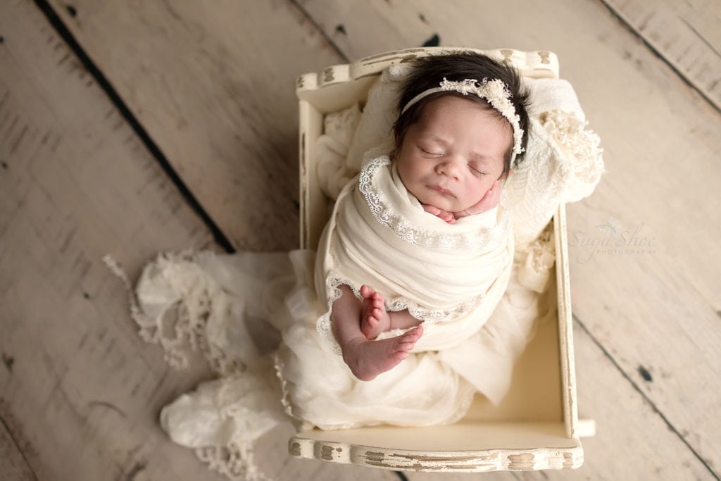 SugaShoc Photography Newborn Photography Bucks County PA Doylestown PA Newborn Photographer Sari Newborn Session baby sleeping in wooden white bed wrapped in white wrap with lace trim and wearing white floral headband