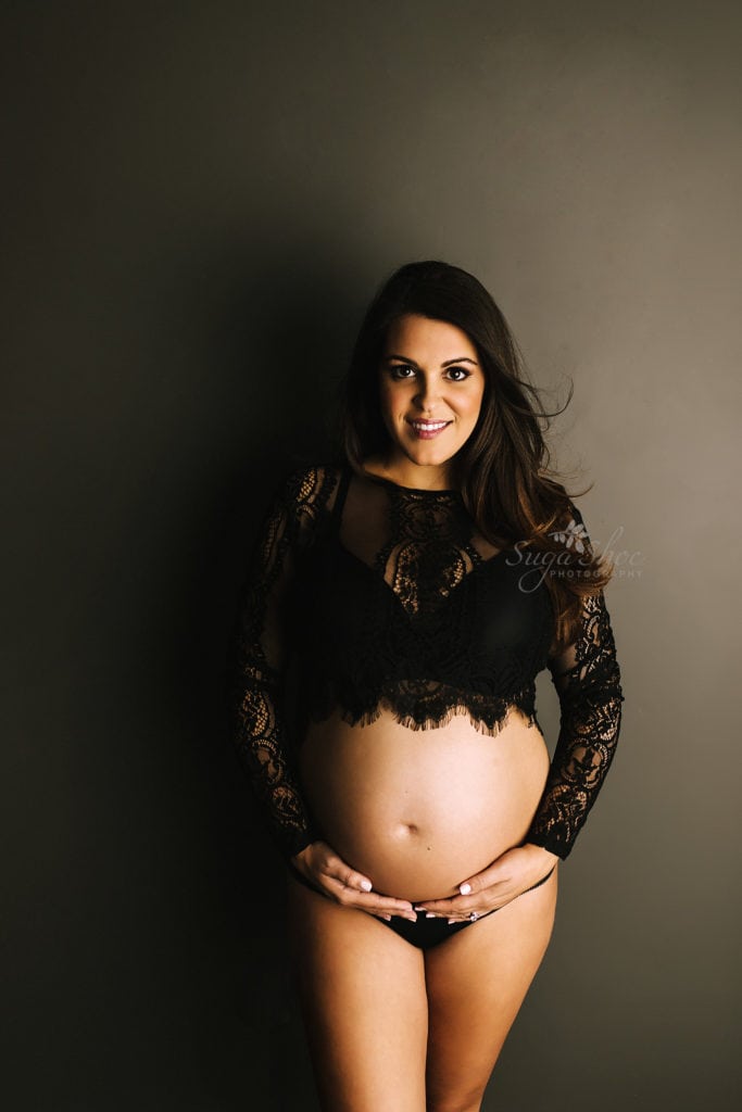 SugaShoc Photography Twin Maternity Session Bucks County PA Doylestown PA Maternity Photographer Maternity pose standing against dark wall wearing black lace top holding belly smiling