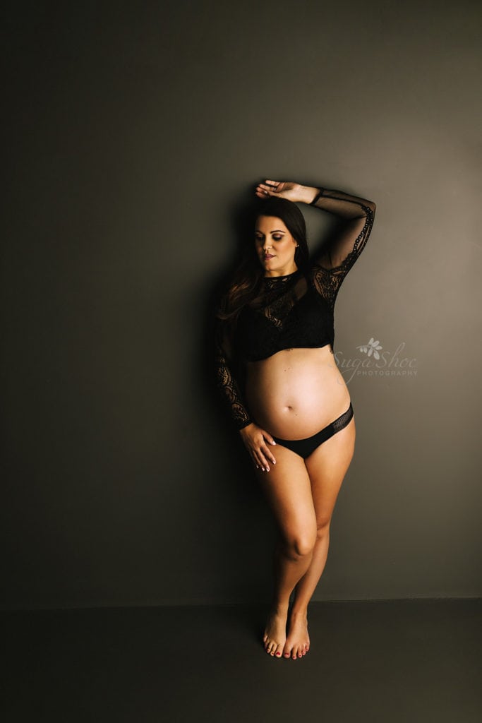 SugaShoc Photography Twin Maternity Session Bucks County PA Doylestown PA Maternity Photographer Maternity pose standing against dark wall wearing black lace top with one arm above head looking down