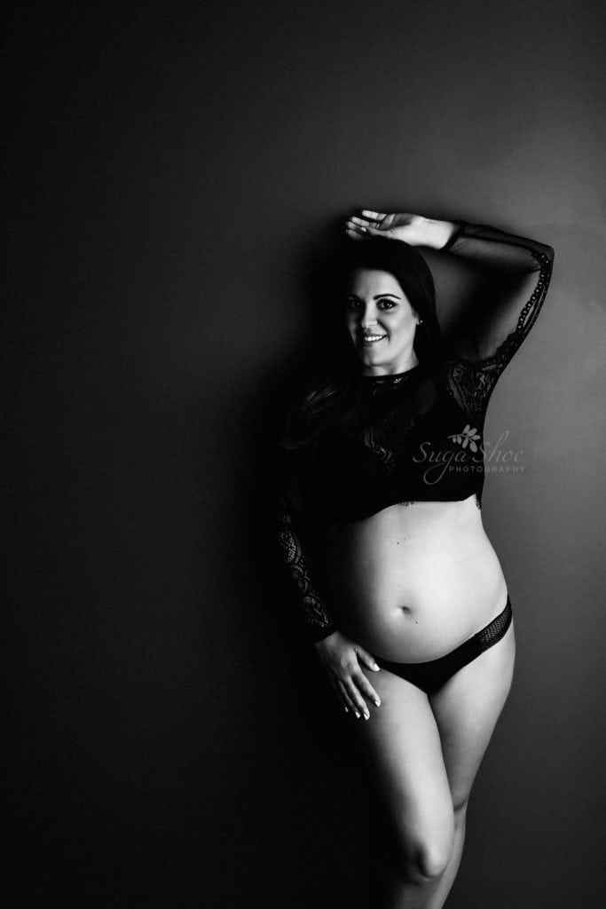 SugaShoc Photography Twin Maternity Session Bucks County PA Doylestown PA Maternity Photographer Maternity pose standing against dark wall wearing black lace top with one arm above head smiling in black-and-white
