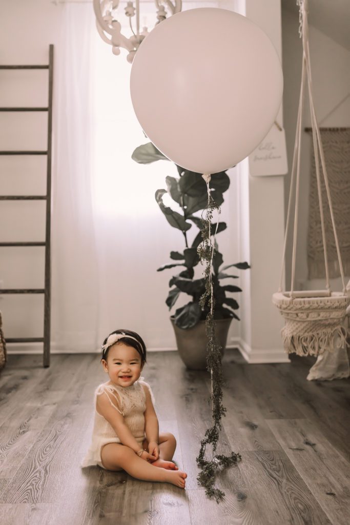 baby girl sitting on floor by window with balloon, plant, and ladder in background