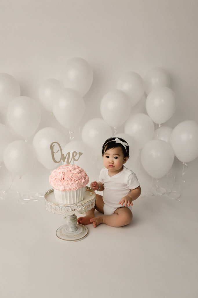 cake smash photo session PA, baby girl against white balloon backdrop with pink cake