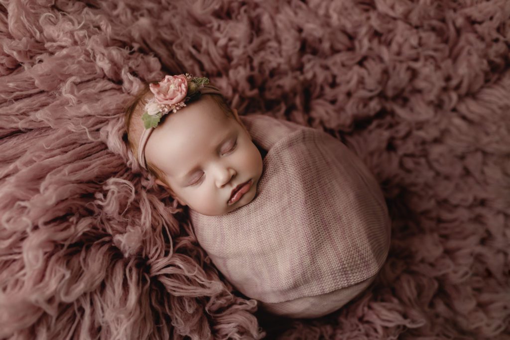 swaddled infant on purple fur throw with floral headband