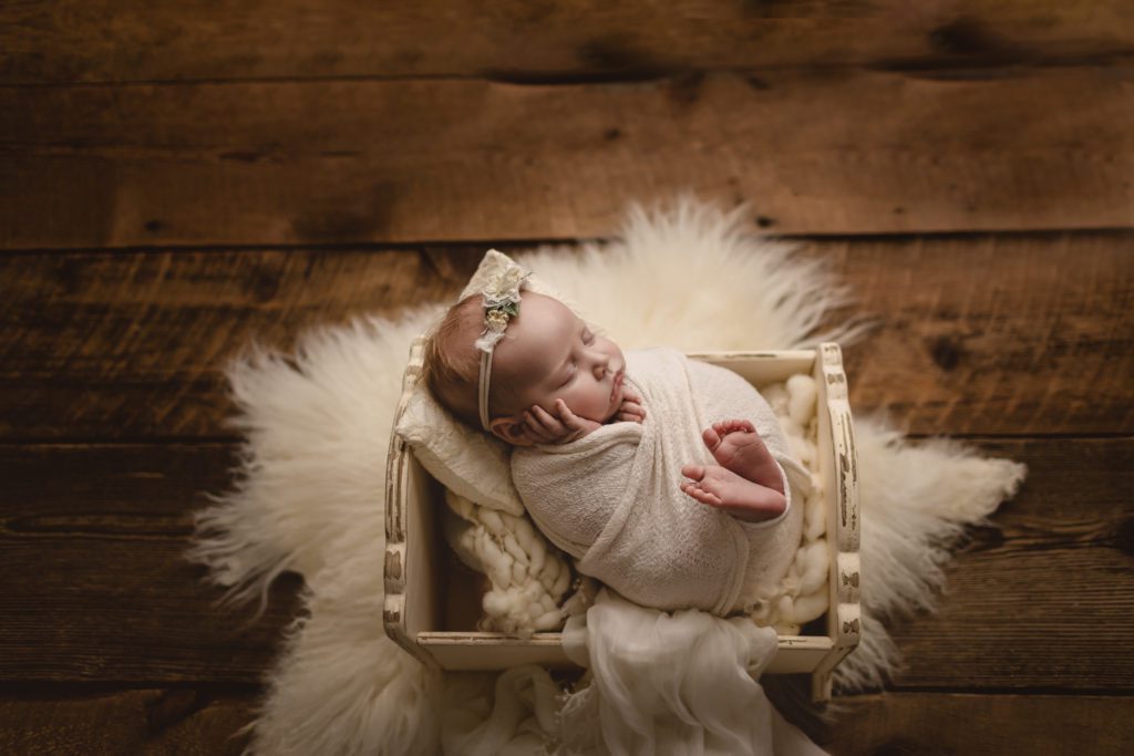 baby in white wrap lying in wooden bed with cream blankets and fur throw