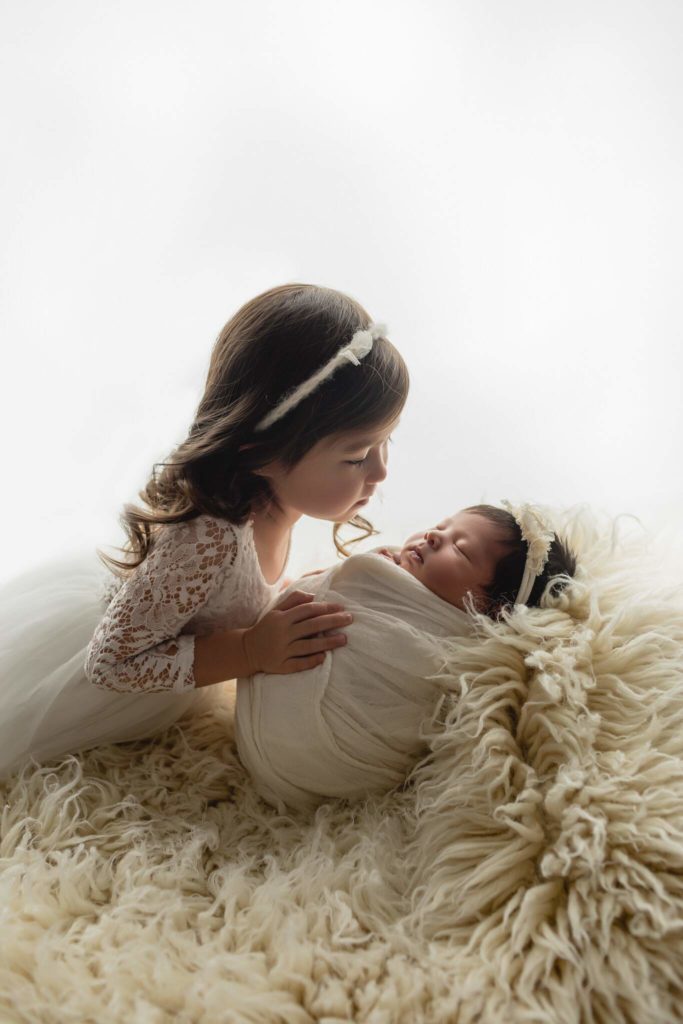 Older sister and newborn sister dressed in white against textured blankets.